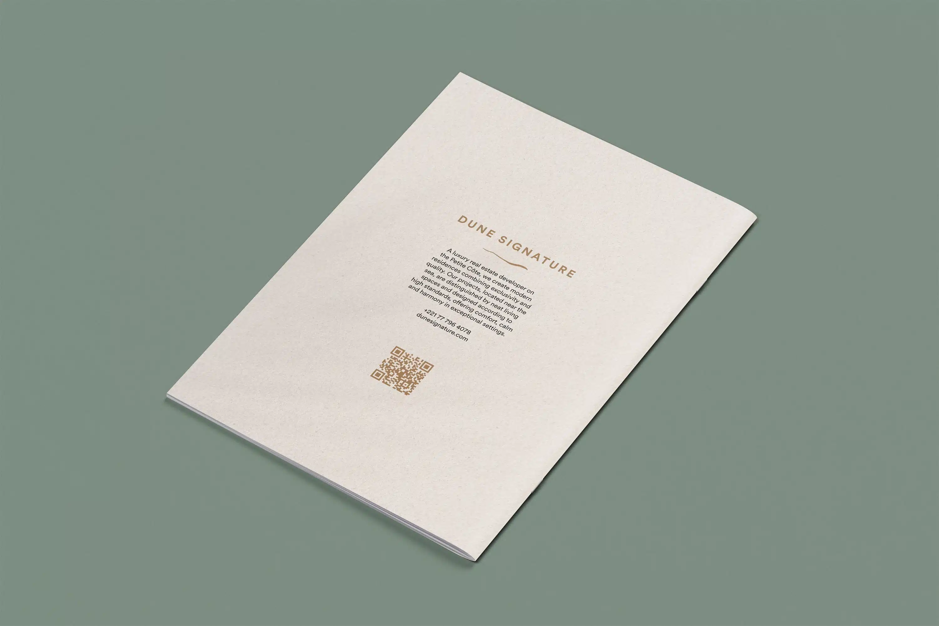 Back cover of the Dune Signature Oasis brochure, featuring the text 'DUNE SIGNATURE' at the top, followed by a paragraph of descriptive text about the company. A gold QR code is visible at the bottom left, and contact information is listed at the bottom right. The brochure is shown at an angle on a grey-green background.