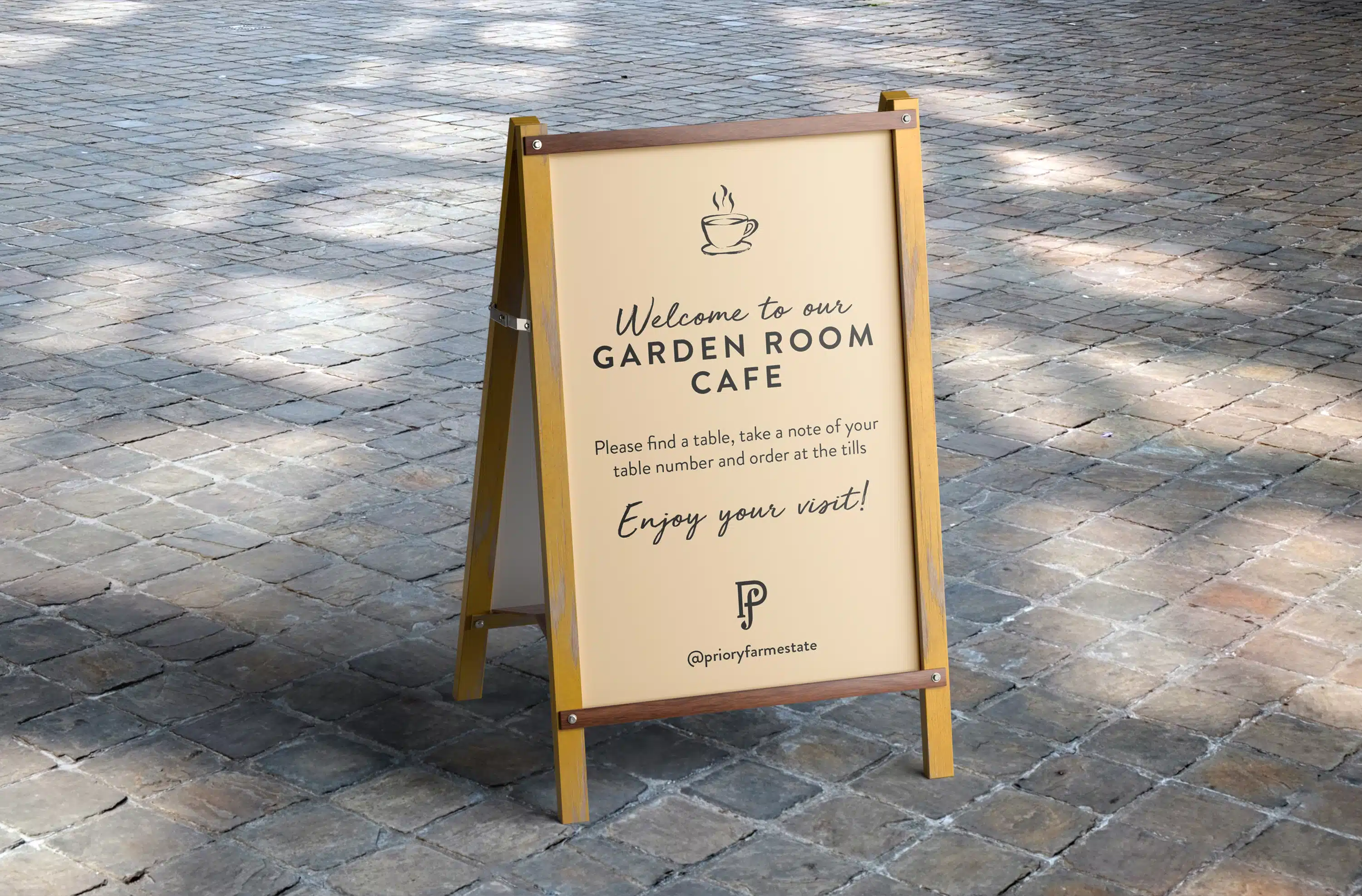 A-frame sign for the Priory Farm Estate Garden Room Café, placed on a paved outdoor surface. The sign features a light-coloured background with a black line-art illustration of a coffee cup at the top, followed by the text 'Welcome to our GARDEN ROOM CAFÉ' in a script font. Below, it reads 'Please find a table, take a note of your table number and order at the tills' and 'Enjoy your visit!' in a sans-serif font. At the bottom, the Priory Farm 'P f' logo and the social media handle '@prioryfarmestate' are displayed.
