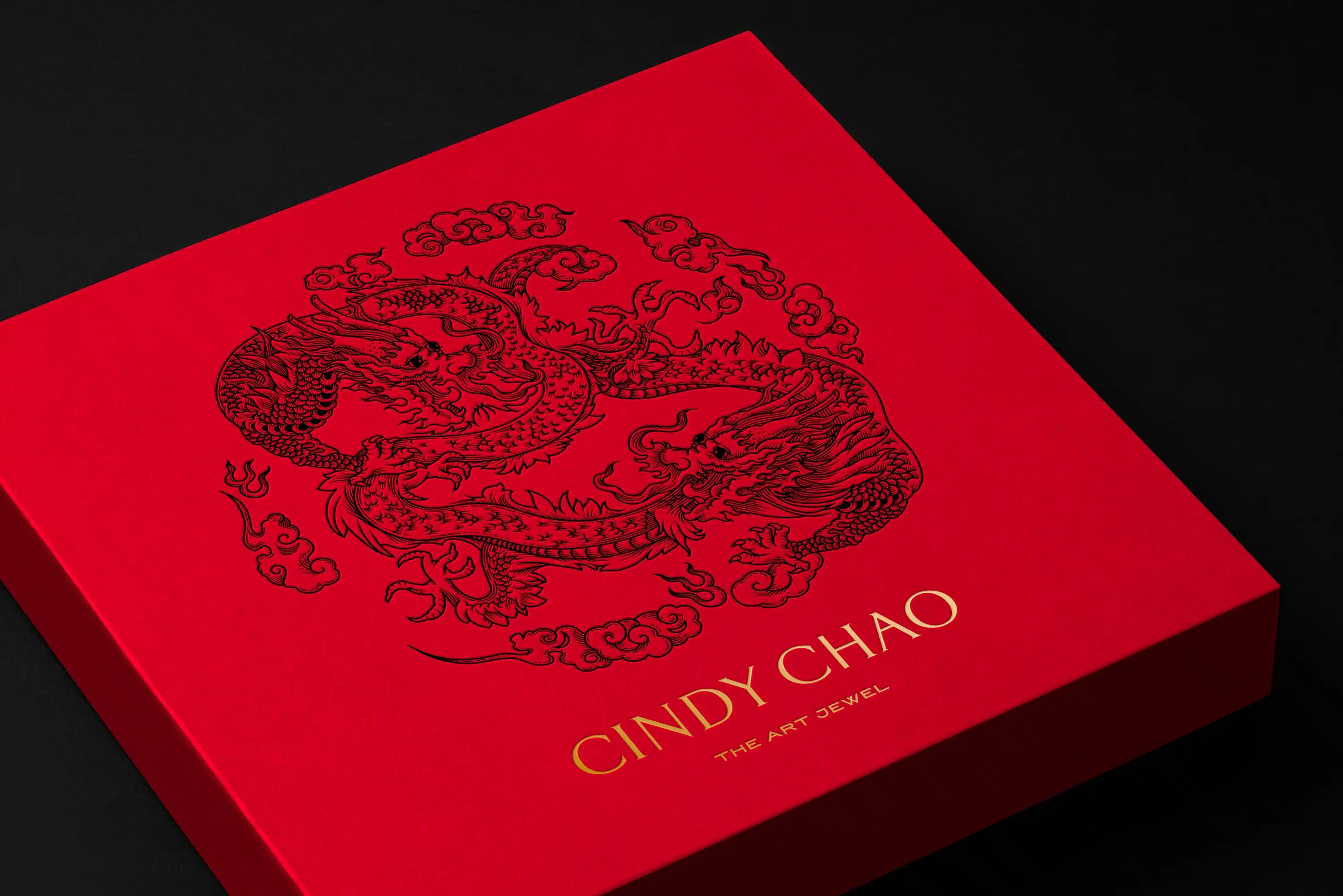 Red square gift box for Cindy Chao's Year of the Dragon celebration, featuring a detailed, contemporary dragon illustration in black against the red background. The box also includes the brand name 'CINDY CHAO' and the tagline 'THE ART JEWEL' in gold lettering at the bottom. The box is presented on a black background, emphasising its vibrant colour and intricate design.